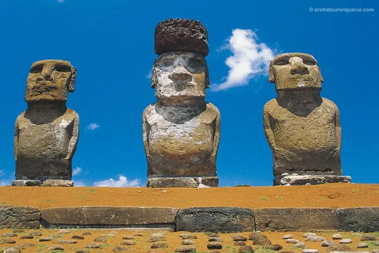 Trois statues Moai, dont l'une porte un "pukao" rouge, sous un ciel bleu à Rapa Nui.